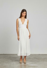 Abito bianco senza maniche con scollatura a V profonda e vita arricciata, caratterizzato da una gonna plissettata fluida che arriva a metà polpaccio.