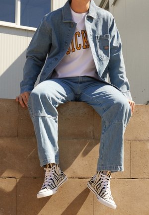 Personne assise sur un mur bas, portant une veste et un pantalon en jean bleu clair, une chemise blanche avec des lettres jaunes, et des baskets montantes à motif zèbre.