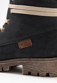 Botín negro de ante con cordones marrón claro, un parche de logo marrón y una suela gruesa y contundente con un diseño texturizado.