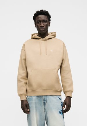Hombre con sudadera beige con bolsillo delantero y jeans azul claro con diseño azul oscuro en el muslo derecho, de pie frente a un fondo liso.