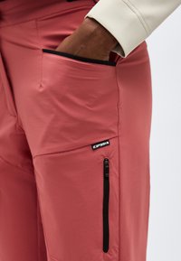 Χέρι στην τσέπη παντελονιού σε σκουριά απόχρωση με μαύρο φερμουάρ και ετικέτα της μάρκας Icepeak στην πλαϊνή τσέπη, που φοράει μανίκι ανοιχτό μπεζ μπουφάν.