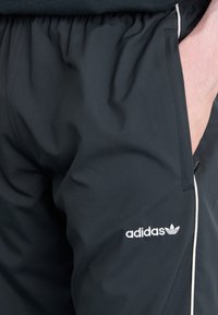Pantaloni sportivi negri cu talie elastică, detaliu din pipă albă și un logo brodat pe piciorul stâng jos. Textura materialului este netedă.