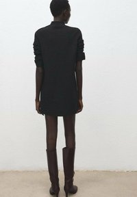 Schwarzes Strickkleid mit übergroßen Ärmeln und hohem Kragen, kombiniert mit kniehohen burgunderroten Stiefeln. Glatte Textur und taillierte Silhouette.