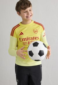 Usmievajúci sa chlapec v žltom futbalovom drese Arsenalu drží čierno-bielu futbalovú loptu pred jednoliatou bielou stenou.