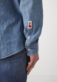 Ljusblå denimskjorta med en slät textur, försedd med en vit etikett med en röd cirkel på ärmen, knäppta manschetter och rundad nederkant.