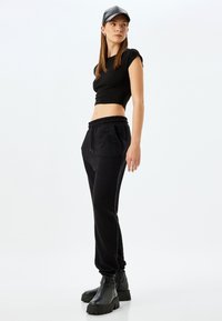Schwarzes, kurzes T-Shirt kombiniert mit schwarzen Sweatpants mit elastischem Bund, seitlichen Taschen und gerafften Bündchen, ergänzt durch schwarze Stiefel.