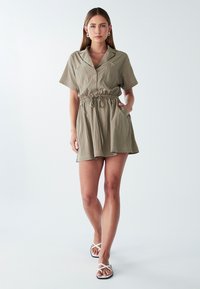 Donna che cammina in avanti indossando un abito verde oliva a maniche corte con vita regolabile e sandali bianchi incrociati su uno sfondo semplice.