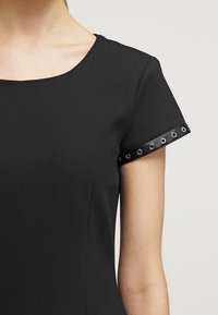 Musta lyhythihainen mekko, jossa on pyöreä pääntie. Ominaisuuksiin kuuluu nahkaranneke, jossa on metallisia niittikoristeita. Sujuva rakenne, vartalonmyötäinen muotoilu.