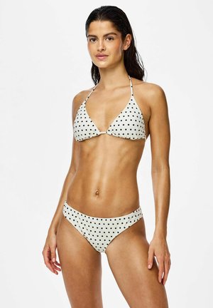 Femeie cu părul udat purtând un bikini alb cu buline negre, stând pe un fundal alb simplu.