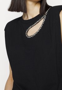 Femme portant un haut noir sans manches avec une découpe en forme de goutte près de l'encolure, soulignée par des ornements en strass brillants.