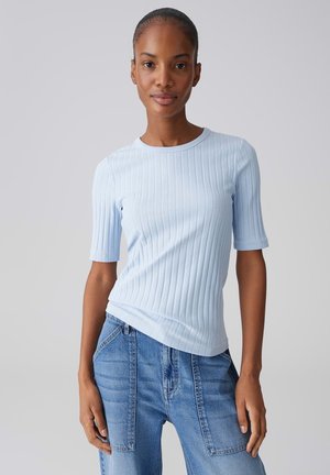 Frau trägt ein hellblaues, geripptes Kurzarmoberteil und hoch taillierte blaue Jeans mit großen Fronttaschen vor einem einfarbigen Hintergrund.