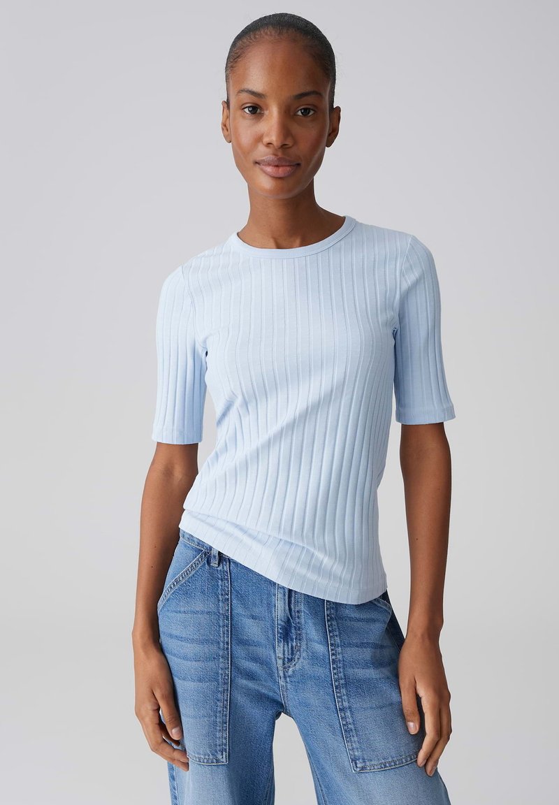 Femme portant un haut à manches courtes côtelé bleu clair et un jean bleu taille haute avec de grandes poches avant, sur un fond uni.