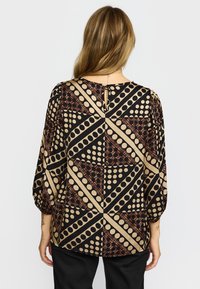 Kvinna med blont hår bär en långärmad blus med ett geometriskt mönster av svarta och beige prickar och former, kombinerad med svarta byxor.