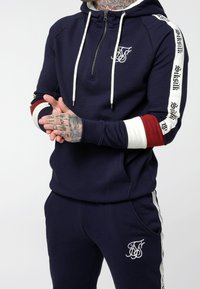 Sudadera de cremallera azul marino con cordones blancos y logo bordado; acentos a rayas en rojo, blanco y negro en las mangas. Combinada con pantalones deportivos a juego.