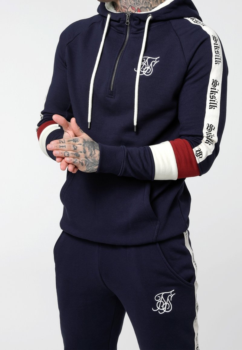 Sudadera de cremallera azul marino con cordones blancos y logo bordado; acentos a rayas en rojo, blanco y negro en las mangas. Combinada con pantalones deportivos a juego.