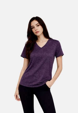 Junge Frau mit langen dunklen Haaren, die ein lila kurzärmeliges V-Ausschnitt-Shirt und schwarze Hosen trägt, steht mit einer Hand in der Tasche.