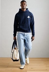 Hanorac bleumarin cu buzunar kangaroo, blugi deschiși la culoare și adidași crem. Poartă o geantă bej și bleumarin. Stil simplu și casual.