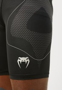 Pantaloni sportivi neri con un design in rete texturizzata, dettagli bianchi e un logo argentato di un serpente. La vestibilità è aderente al corpo.