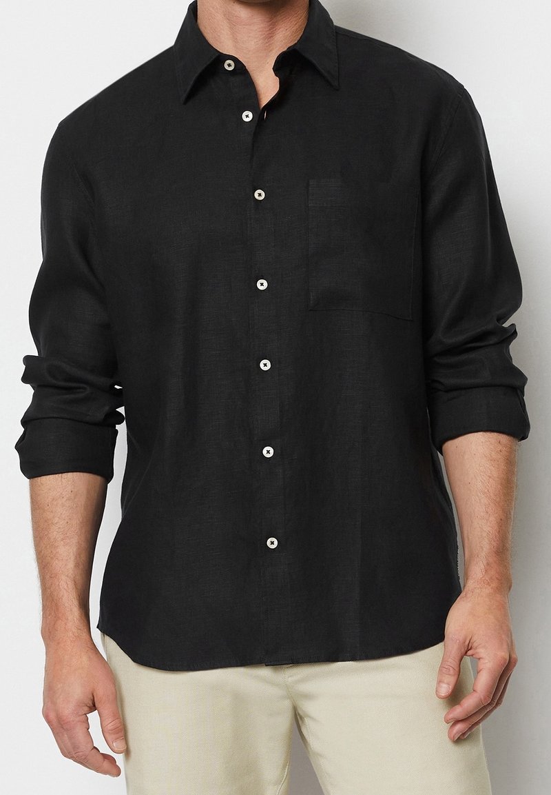 Camisa de lino negra con botones, con bolsillo, mangas enrolladas y botones blancos contrastantes. Textura suave con un corte relajado y dobladillo curvado.