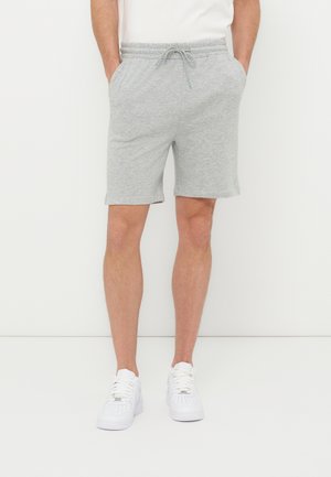 Άνδρας που φοράει ανοιχτό γκρι σορτς με κορδόνια και λευκά αθλητικά παπούτσια, στέκεται με τα χέρια στις τσέπες μπροστά σε απλό λευκό φόντο.