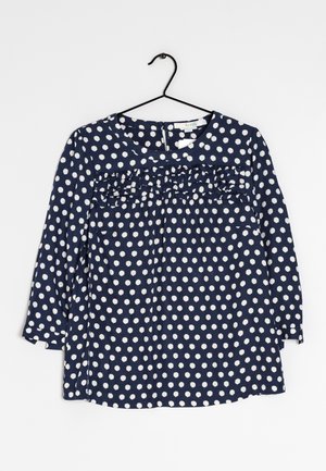Navy blue long-sleeve blouse with white polka dots, round neckline, ruffle detail across chest, hanging on black hanger.