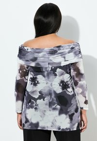 Femme aux longs cheveux noirs portant un haut à épaules dénudées à manches longues avec un motif floral abstrait noir et blanc, faisant face à l'arrière.