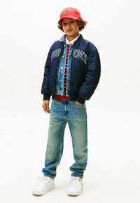 Marine bomberjakke med hvidt skrift, denimvest, rødternet skjorte, lyseblå jeans og hvide sneakers, toppet med en rød kasket.