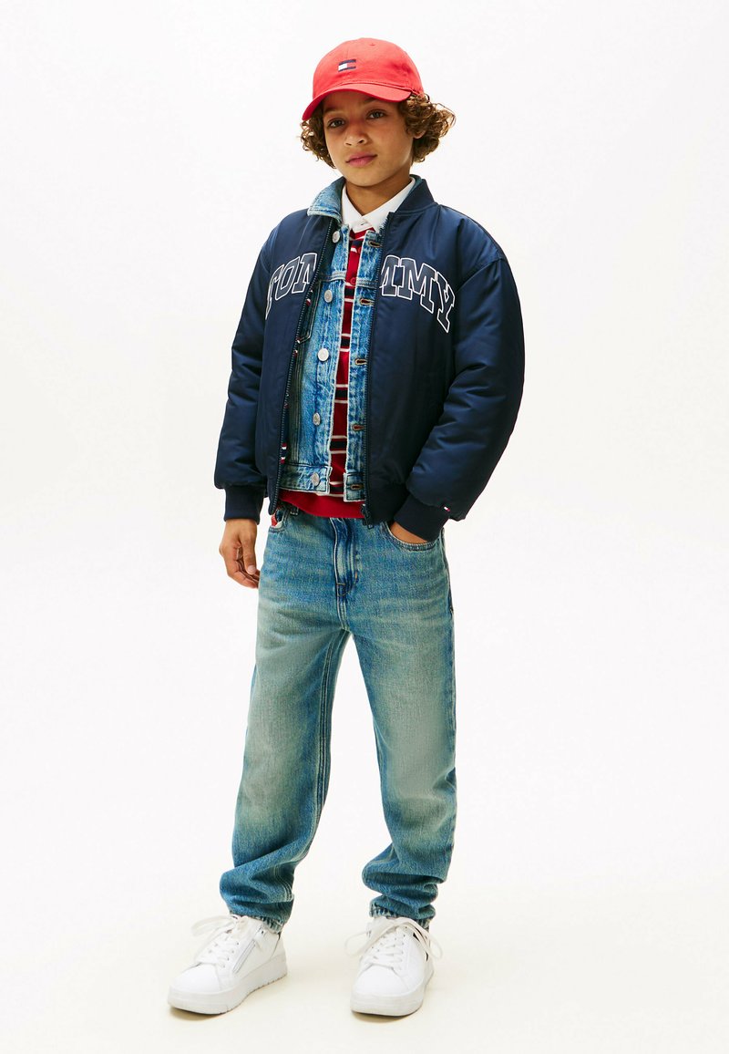 Marine bomberjakke med hvidt skrift, denimvest, rødternet skjorte, lyseblå jeans og hvide sneakers, toppet med en rød kasket.