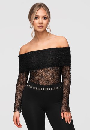 Femme aux cheveux blonds portant un top en dentelle noire épaules dénudées et un pantalon noir avec des clous argentés, debout devant un fond clair uni.