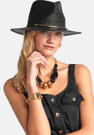 Femme portant un chapeau tissé noir, un top noir sans manches avec des boutons dorés, un collier épais noir et or, et un bracelet en or, posant avec la main près du visage.