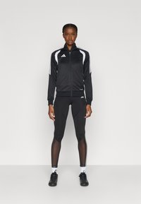 Μαύρο αθλητικό μπουφάν με λευκές ρίγες, μπροστινό φερμουάρ, σε συνδυασμό με μαύρο κολάν που διαθέτει διακοσμητικά στοιχεία από δίχτυ και υφή στο υλικό, φορεμένα με μαύρα παπούτσια.