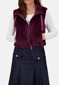 Bordeauxfarbene Kunstfellweste mit Reißverschluss, zwei vorderen Taschen und Bandakzenten, kombiniert mit einem dunklen Jeansrock mit Knopfdetails.