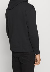 Černá bavlněná mikina s volným střihem, velkou kapucí, žebrovanými manžetami a lemem. Hladký povrch, bez viditelných vzorů či log. 