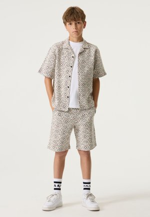 Chemise à manches courtes à motifs et short assorti en tissu léger, avec un design géométrique aux tons beige et gris. T-shirt blanc en dessous.
