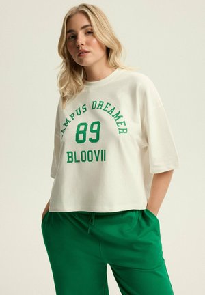 Biała krótka koszulka z zielonym napisem "CAMPUS DREAMER 89 BLOOVII" w zestawie z zielonymi dresami. Miękka bawełniana tkanina, luźny krój.