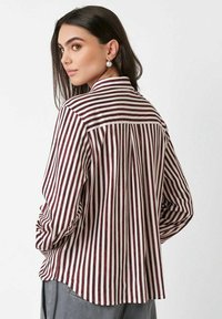 Young woman with long dark hair wears pearl earrings and a loose white shirt with vertical dark red stripes, facing backward, head turned right.
