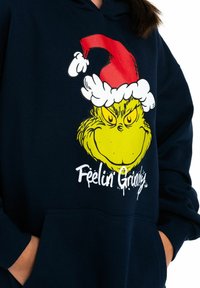 Persona che indossa un hoodie blu navy con la faccia del Grinch gialla, un cappello di Babbo Natale rosso e la scritta "Feelin Grinchy" sul davanti.