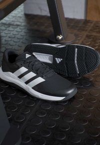 Paire de chaussures de sport Adidas noir et blanc sur un sol de gym texturé, une chaussure debout et l'autre montrant la semelle.