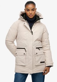 Beżowa ocieplana parka z kapturem, zapinana na suwak z przodu, z dwoma przednimi kieszeniami. Posiada czarne akcenty suwakowe i miękką teksturę.