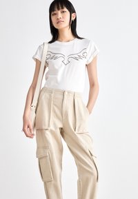 Weißes T-Shirt mit einem grafischen schwarzen Umriss, kombiniert mit hellbeigen Cargo-Hosen mit Seitentaschen und lockerer Passform.