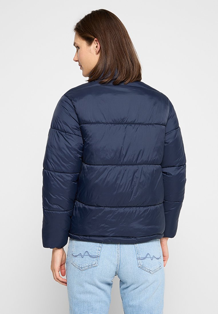 Chaqueta acolchada azul marino de corte recto, con secciones cosidas y una textura suave y brillante. Vista posterior, llevada sobre unos vaqueros de corte claro.