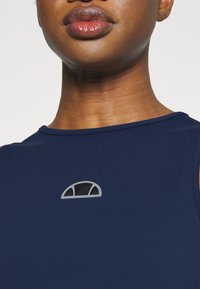 Granatowy top bez rękawów z okrągłym dekoltem, z szarym logo w kształcie półokręgu z wyciętymi detalami. Gładka faktura tkaniny.