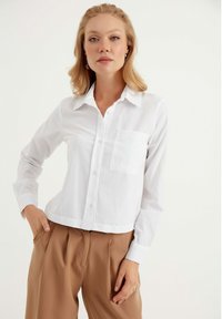 Witte button-up blouse met een kraag, enkel borstzakje en lange mouwen. Gecombineerd met losse, tan kleurige broeken. Glad stofgevoel.