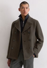 Oliwkowy wełniany płaszcz typu peacoat z podwójnym zapięciem, dużymi czarnymi guzikami i szerokimi klapami, w zestawie z czarnym swetrem zapinanym na zamek.