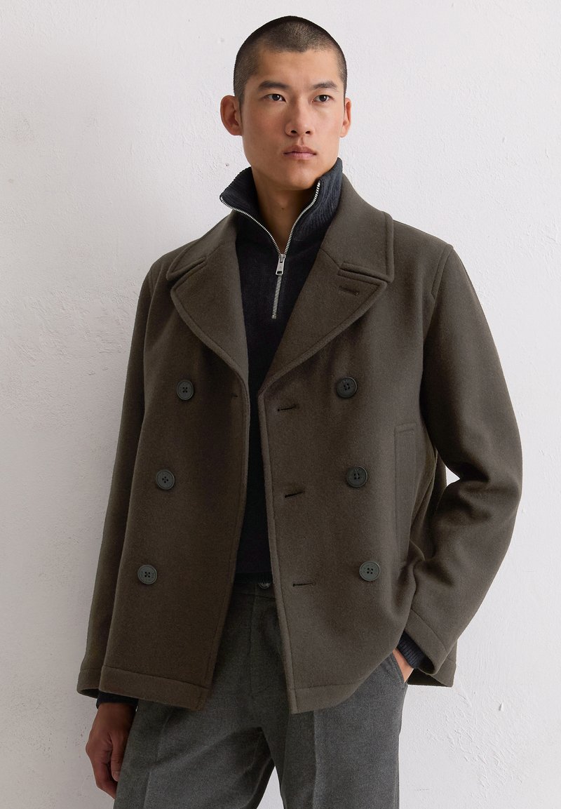 Oliwkowy wełniany płaszcz typu peacoat z podwójnym zapięciem, dużymi czarnymi guzikami i szerokimi klapami, w zestawie z czarnym swetrem zapinanym na zamek.