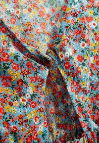 Tissu bleu avec un petit imprimé floral coloré en rouge, jaune, blanc et vert, présentant des plis rassemblés et torsadés.