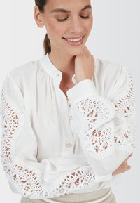 Weiße Bluse mit bestickten Spitzenärmeln, die über ausgefallene Ausschnittmuster verfügt, mit Knopfleiste und aus einem weichen, leichten Stoff gefertigt.