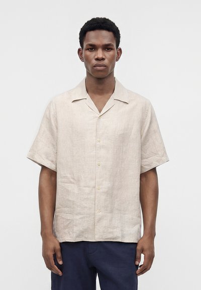 Homme portant une chemise beige à manches courtes boutonnée et un pantalon bleu marine, debout devant un fond clair uni, regardant droit devant.