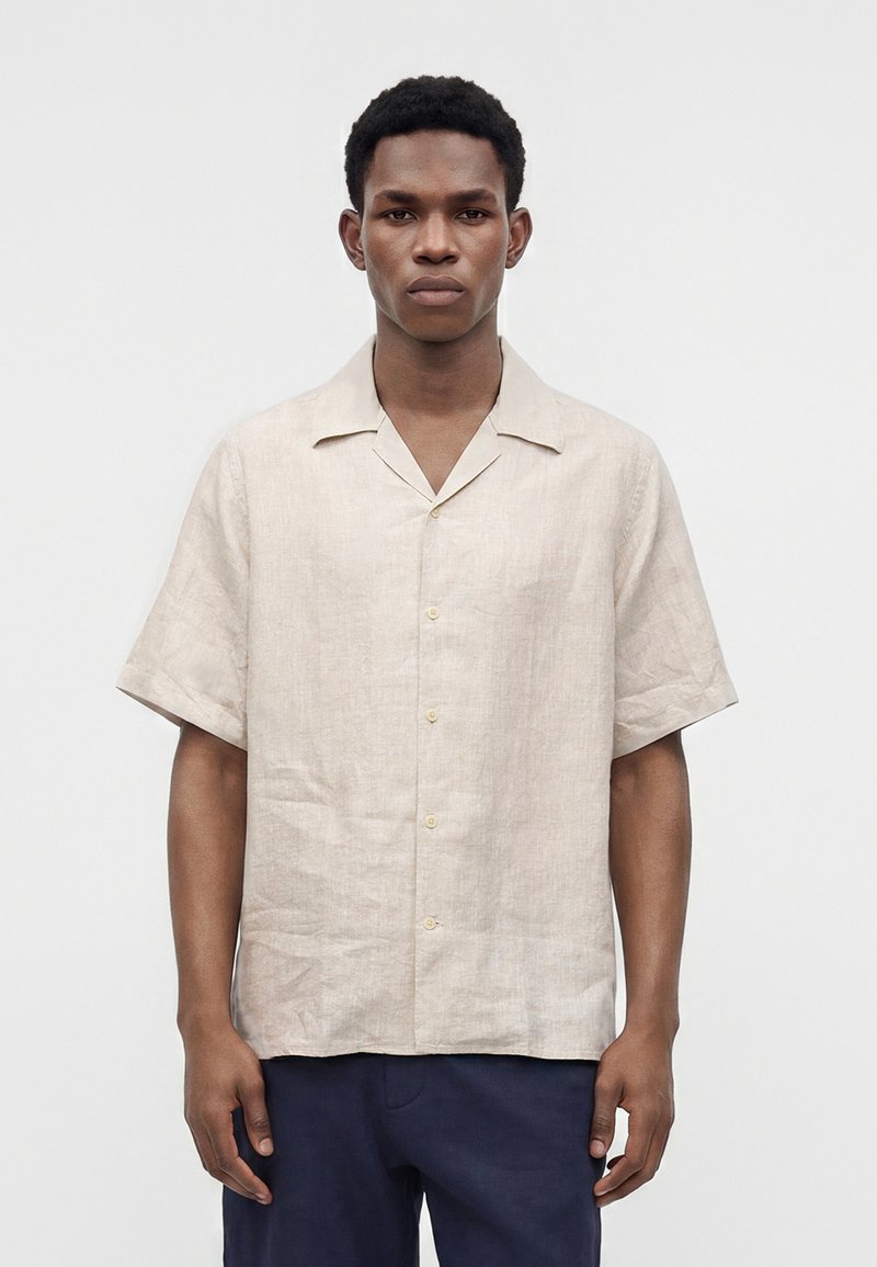 Homme portant une chemise beige à manches courtes boutonnée et un pantalon bleu marine, debout devant un fond clair uni, regardant droit devant.