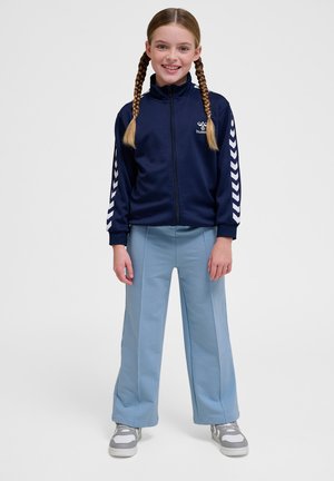 Marineblauwe ritsjack met witte chevronstrepen op de mouwen, gecombineerd met lichtblauwe wijde broek en grijze sneakers.